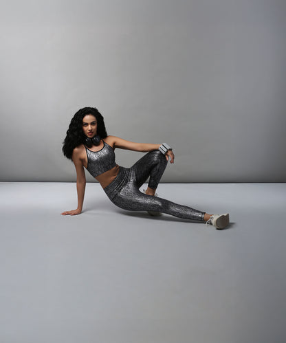 A woman models the Black Silver Shimmer Leggings, sitting on the floor in gray athletic wear against a plain gray background.