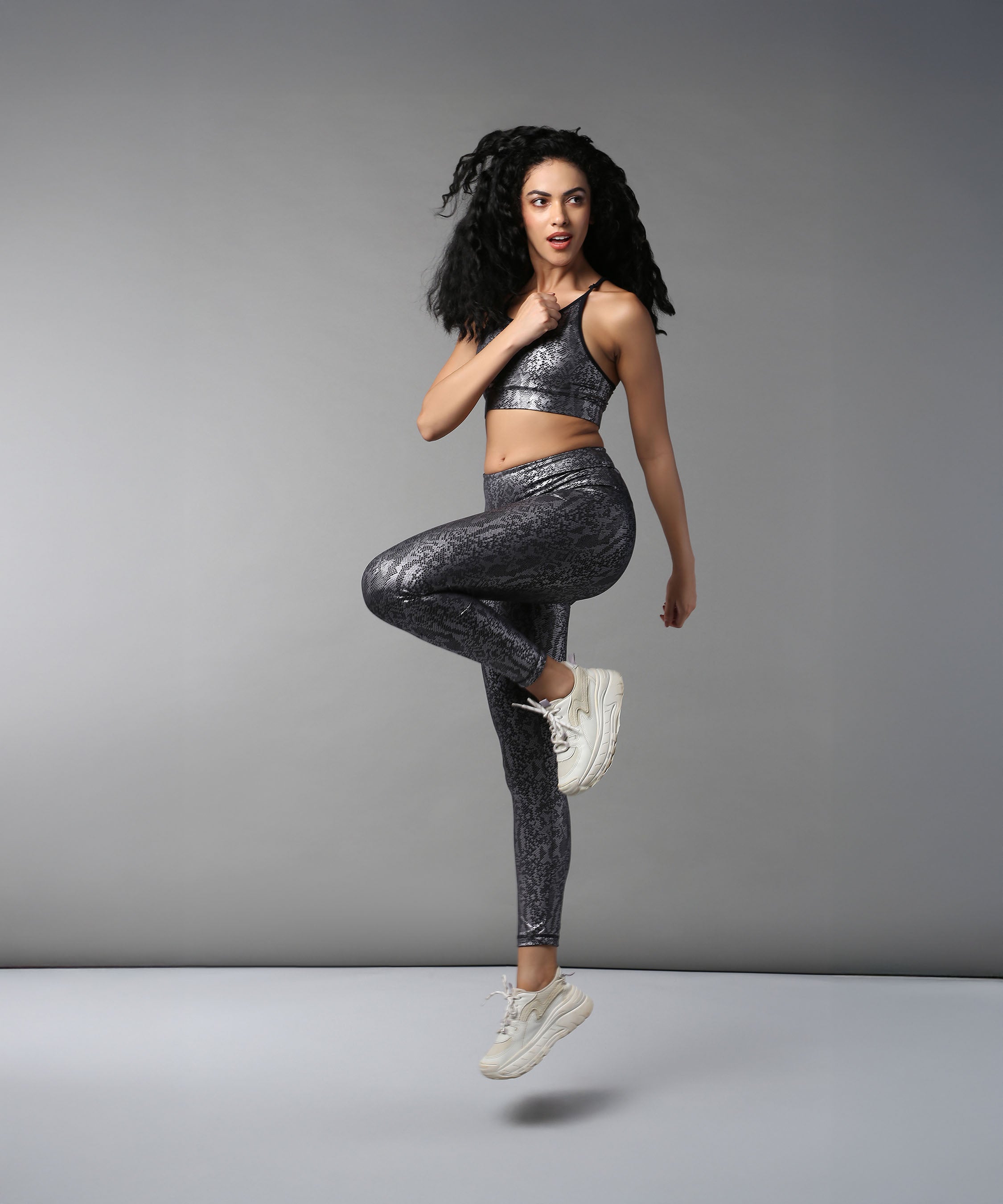 A woman in athletic wear jumps with one knee raised against a grey background, showcasing Black Silver Shimmer Leggings crafted from high-performance fabric.
