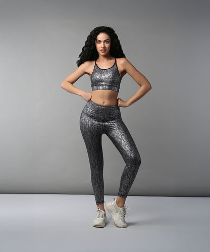 A woman wears Black Silver Shimmer Leggings, made from high-performance fabric, and white sneakers as she stands with hands on hips against a plain gray backdrop.