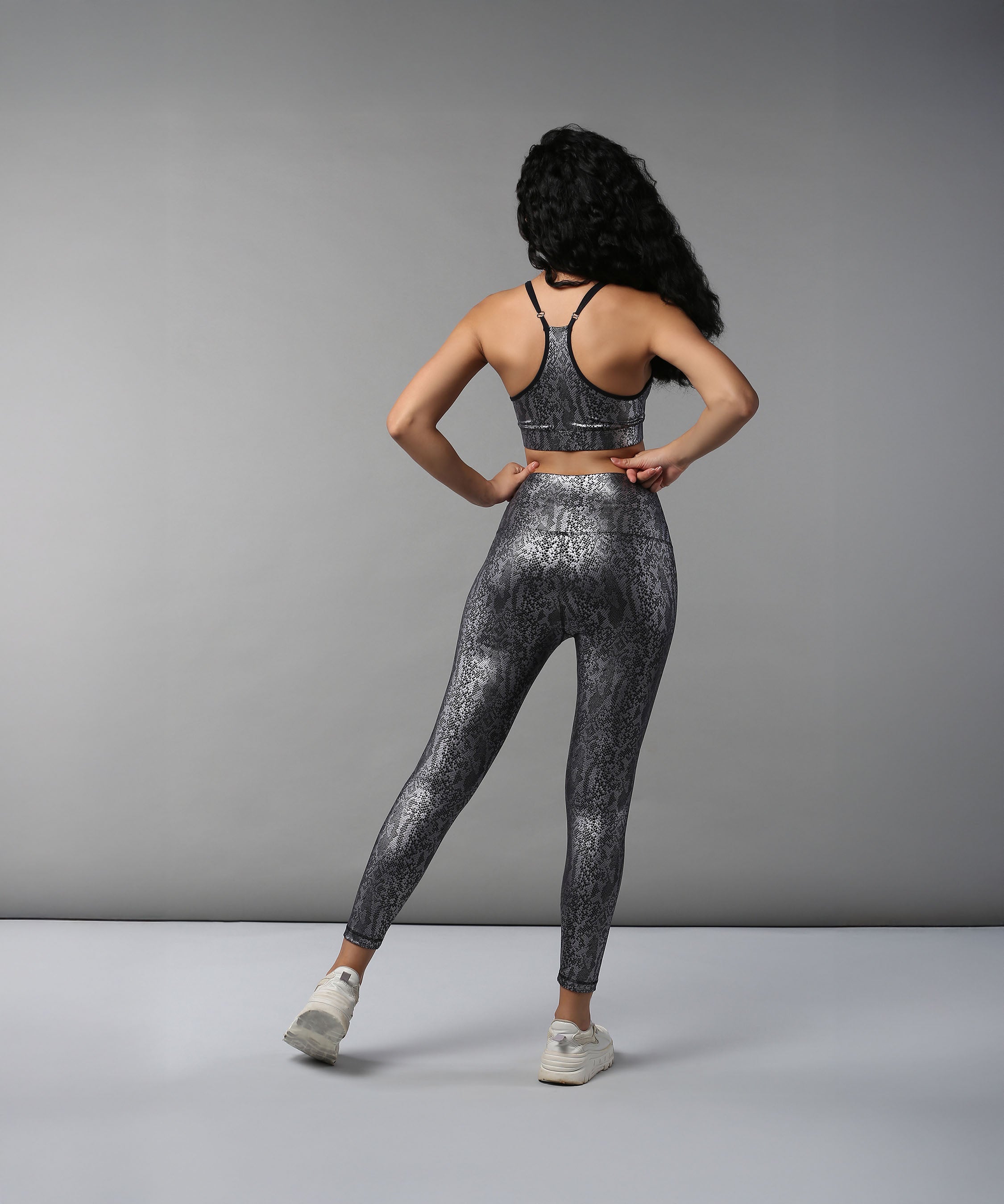 A woman stands in a studio setting wearing the Black Silver Shimmer Leggings with a coordinating snakeskin-patterned sports bra, facing away.