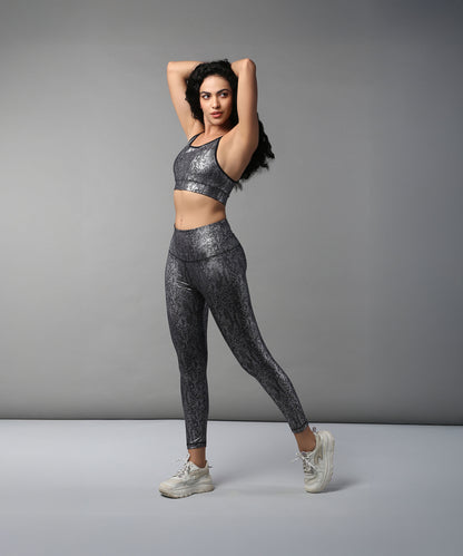 Woman wears Black Silver Shimmer Leggings made from high-performance fabric, posing with arms raised and balancing on one leg against a plain background.
