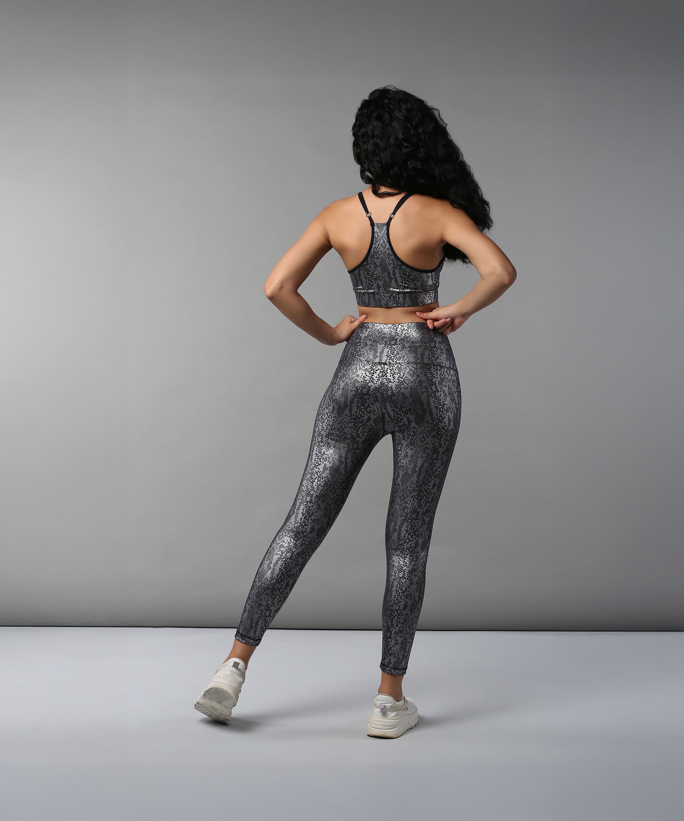 Woman with curly hair faces away from the camera in front of a plain gray backdrop, wearing the Black Silver Shimmer Print Sports Bra and matching activewear.