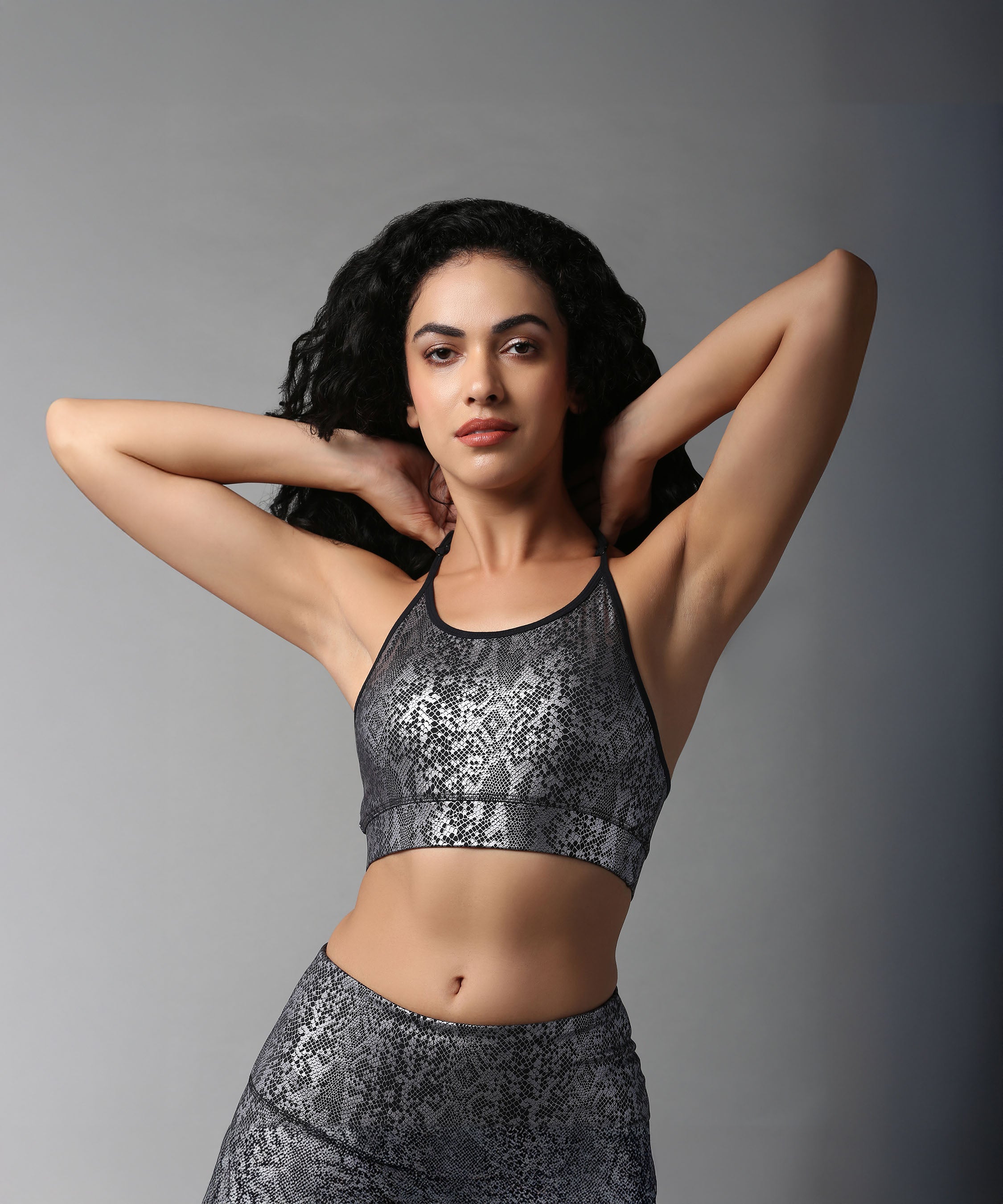 Woman with curly hair poses with hands behind her head, wearing the Black Silver Shimmer Print Sports Bra and matching workout outfit against a gray background.