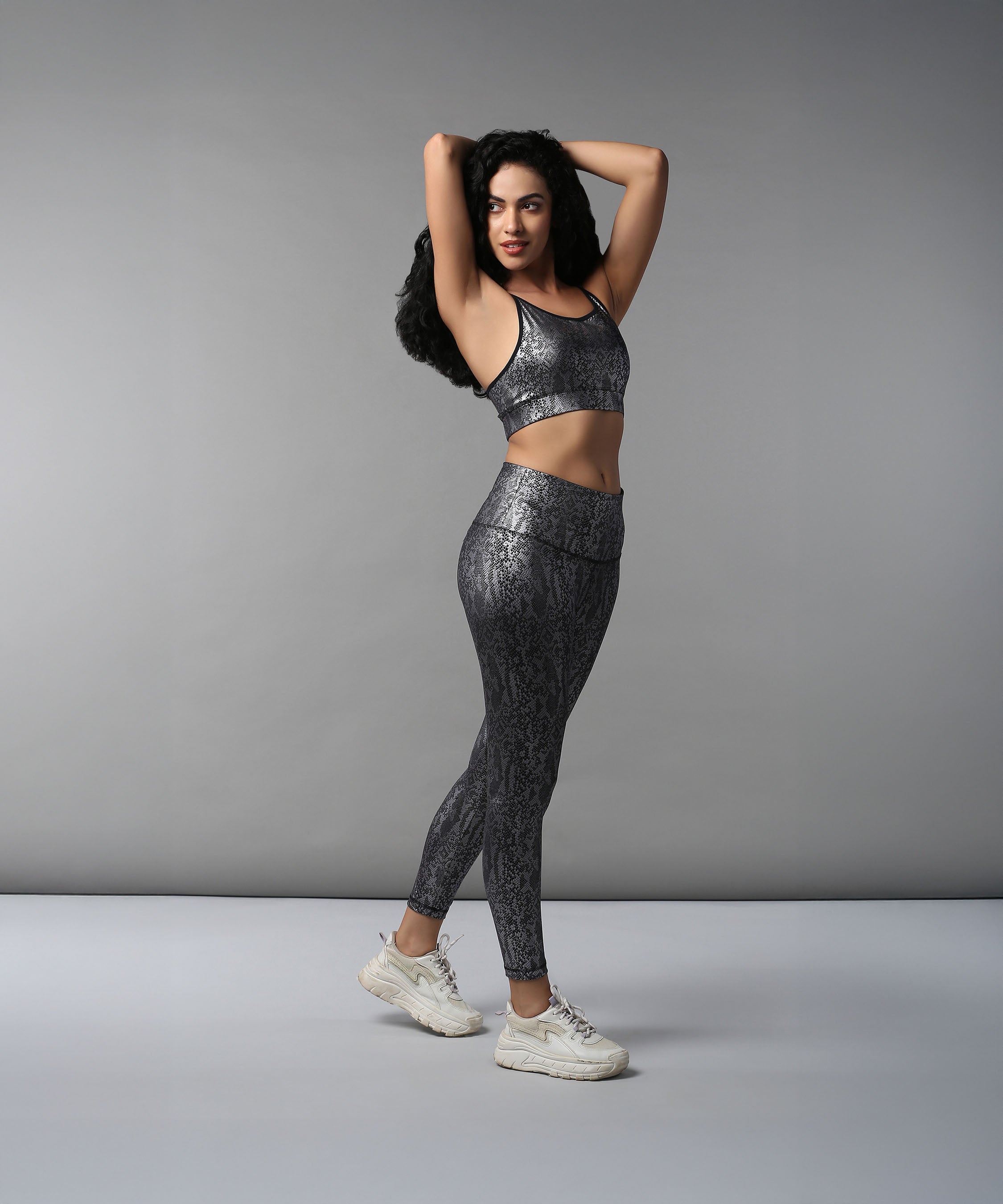 Woman poses confidently against a plain gray background, wearing the Black Silver Shimmer Print Sports Bra with shiny patterned workout clothes and white sneakers.