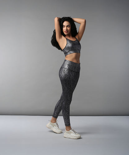 Woman poses confidently against a plain gray background, wearing the Black Silver Shimmer Print Sports Bra with shiny patterned workout clothes and white sneakers.