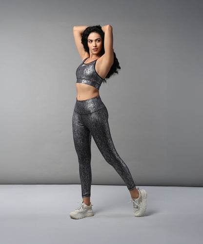 Woman wears the Black Silver Shimmer Print Sports Bra with activewear, posing with arms raised and standing on one leg against a plain background.