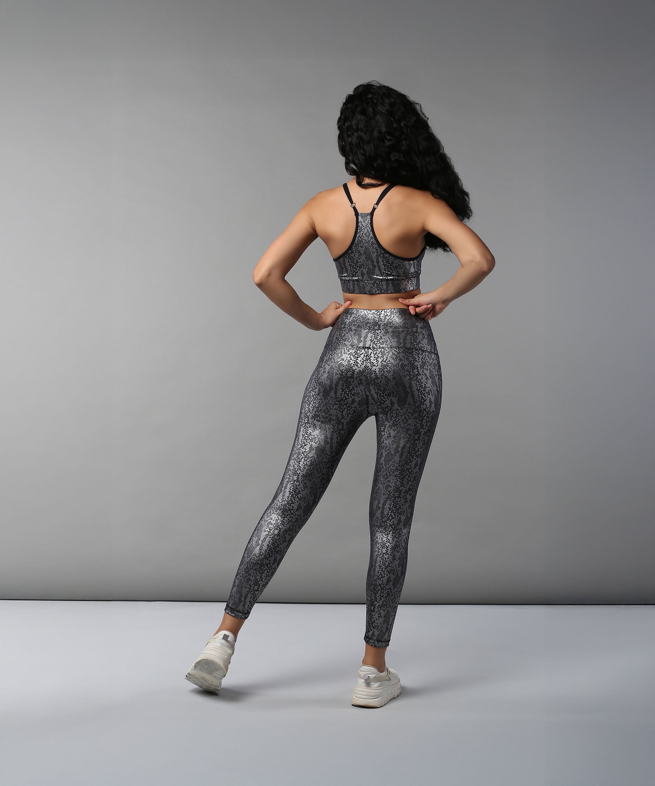Woman with long curly hair stands facing away, wearing the Black Silver Shimmer Print Sports Bra and high waisted Leggings set.