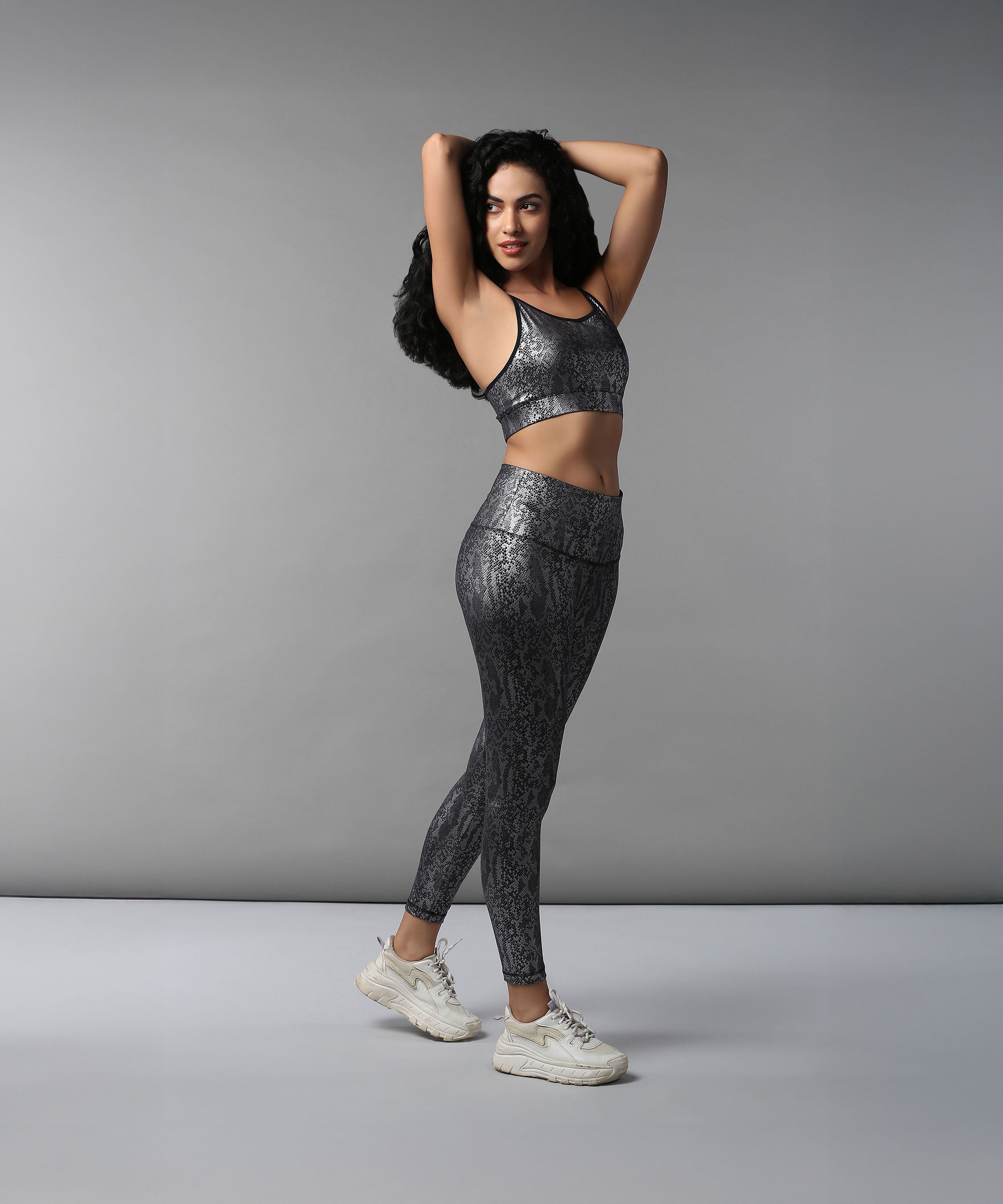 Woman in Black Silver Shimmer Print Sports Bra and Leggings poses confidently in white sneakers against a gray background, highlighting the comfort of the moisture-wicking fabric.