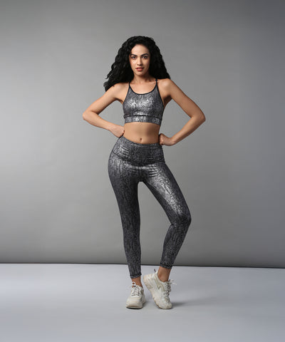 A woman stands with hands on hips against a plain gray background, wearing the Black Silver Shimmer Print Sports Bra and Leggings, highlighting the breathable, moisture-wicking fabric.