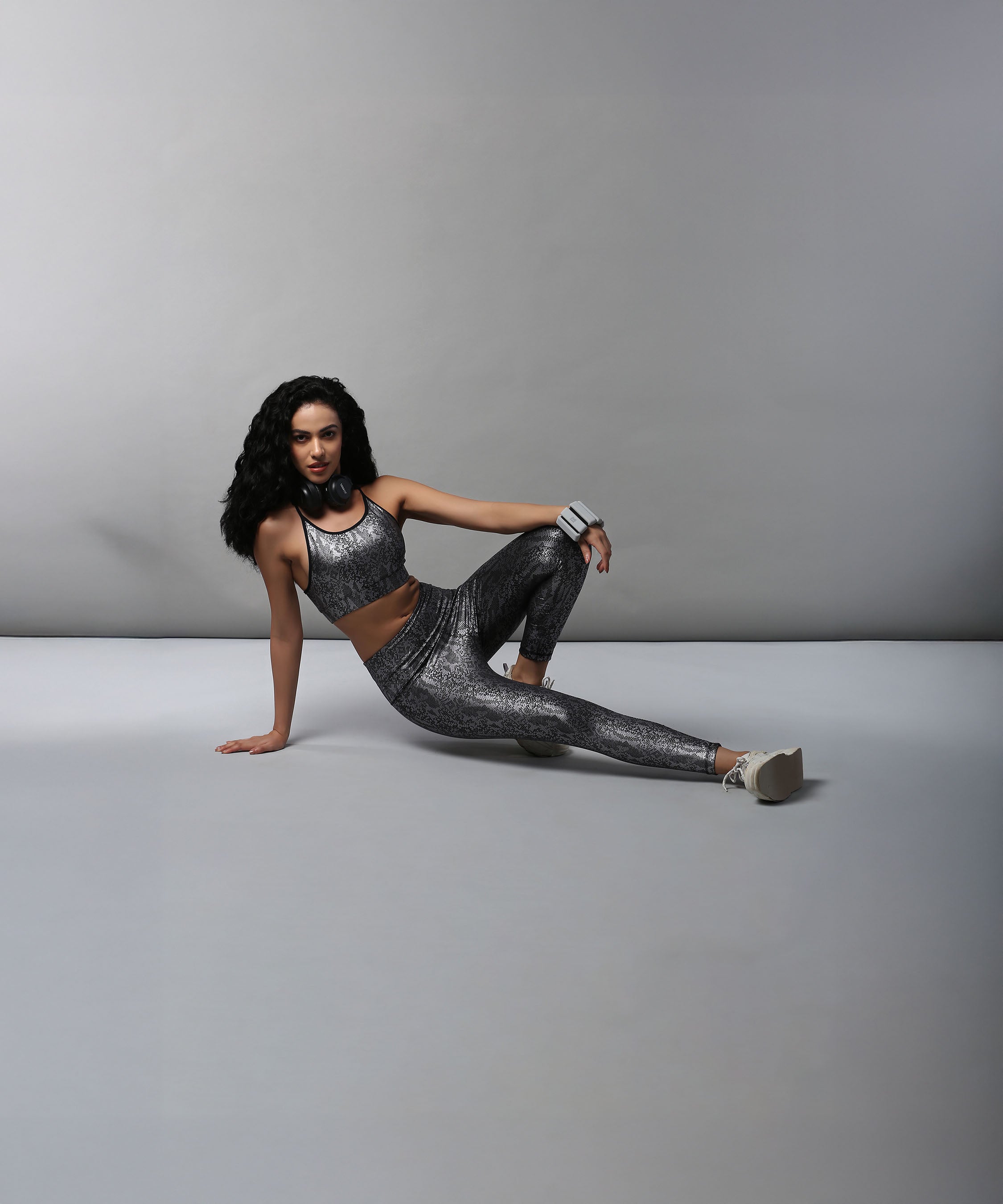 A woman with long dark hair poses on the floor in the Black Silver Shimmer Print Sports Bra and Leggings, featuring moisture-wicking fabric, against a plain gray background.