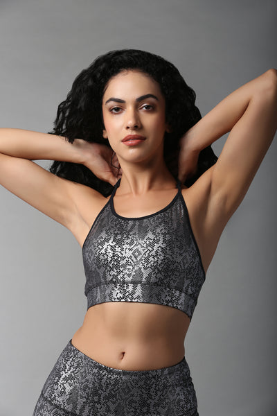 A woman with curly hair poses with her hands behind her head against a gray background, wearing the Black Silver Shimmer Print Sports Bra and high-waisted leggings.