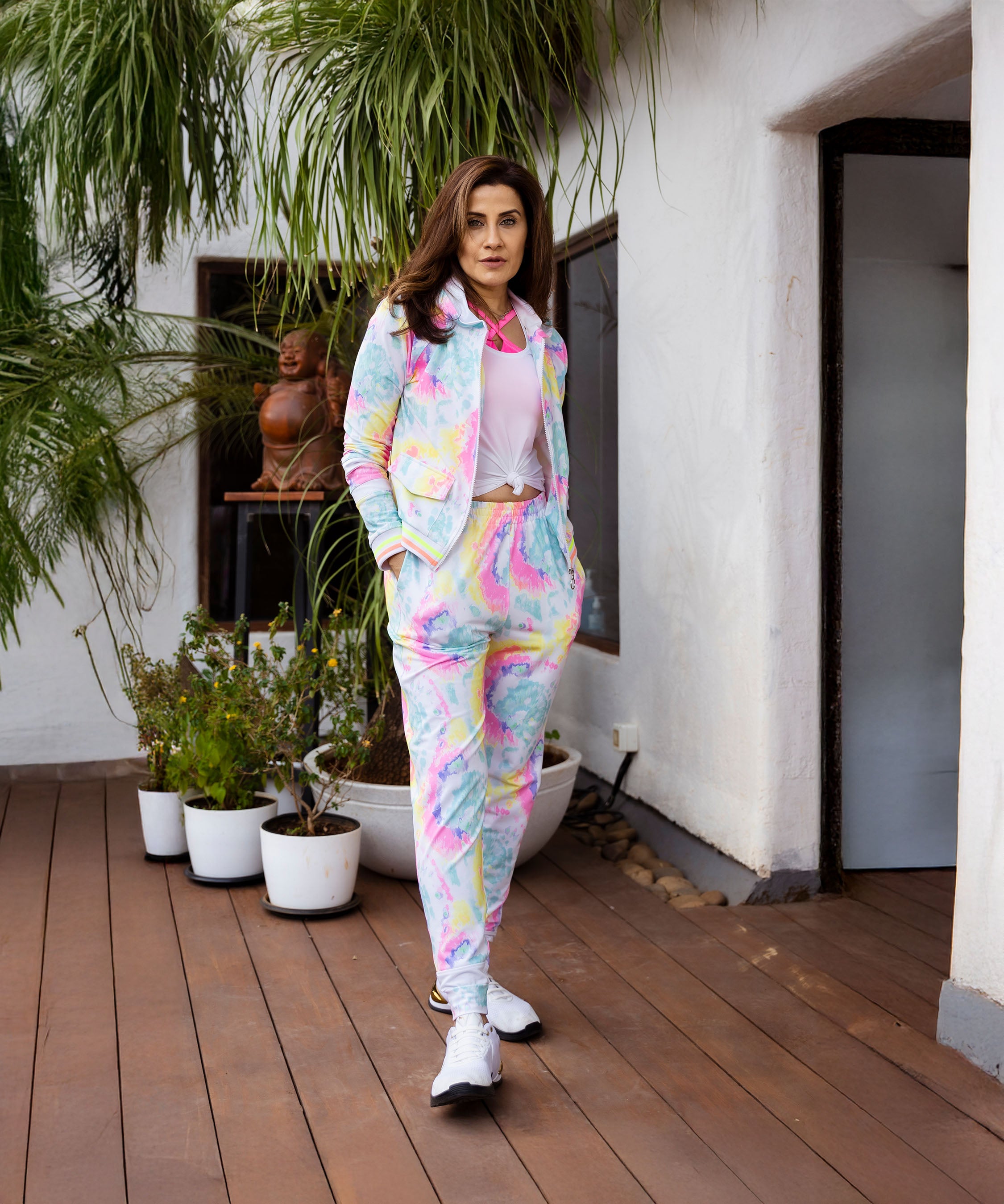 A woman stands on a deck with potted plants and white walls, wearing the Multicolour Neon Jacket & Joggers Set featuring a vibrant poly-blend fabric and casual cargo pockets.