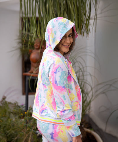 A woman smiles over her shoulder, standing by greenery and white walls, wearing the Multicolour Neon Jacket & Joggers Set. The vibrant poly blend fabric adds comfort and style to her cheerful look.