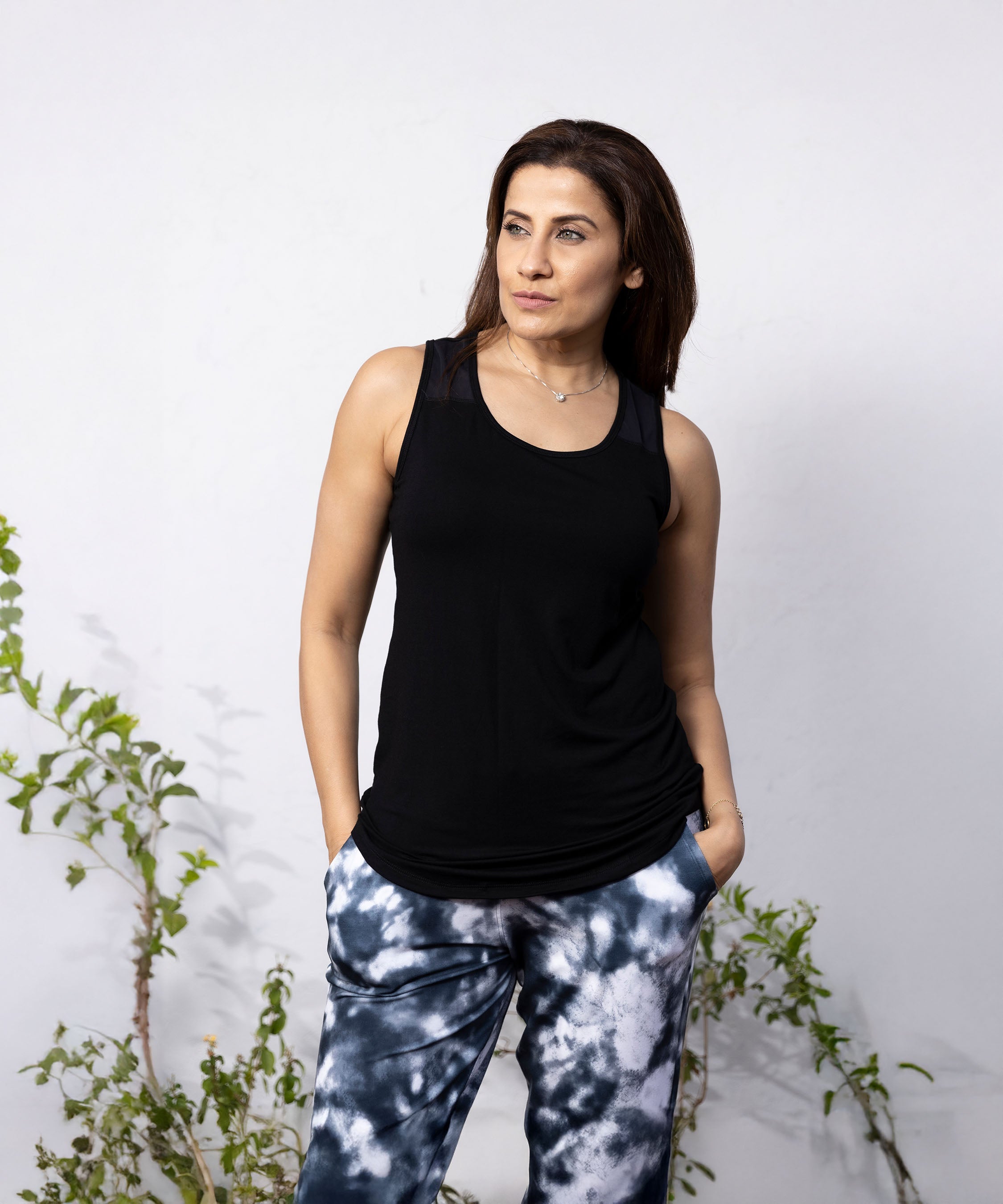 A woman wears the 'Pilates Has Your Back' Printed Racer Black Top and tie-dye pants, standing with hands in her pockets and looking to the side.