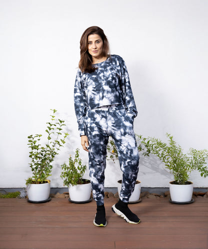 A woman wearing a Grey and White Sweatshirt-Joggers Set stands on a wooden deck with potted plants against a white wall.