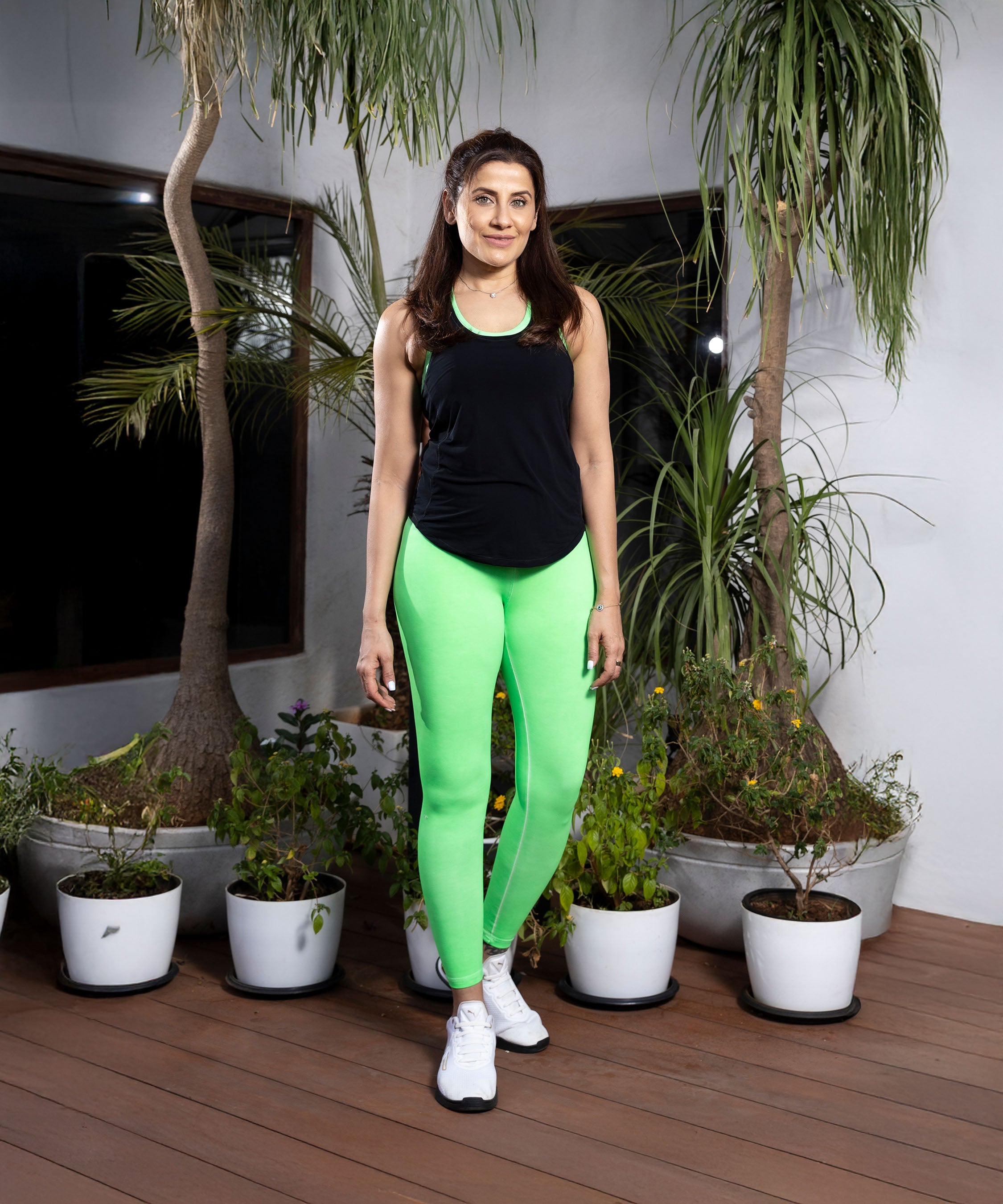 Smiling at the camera, a woman stands indoors near potted plants wearing Neon Green Leggings with a high-rise waistband and a black tank top.