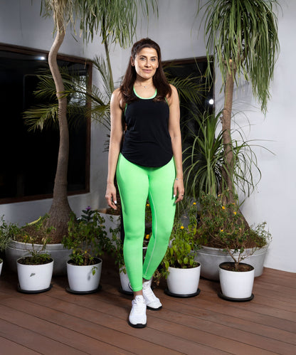Smiling at the camera, a woman stands indoors near potted plants wearing Neon Green Leggings with a high-rise waistband and a black tank top.