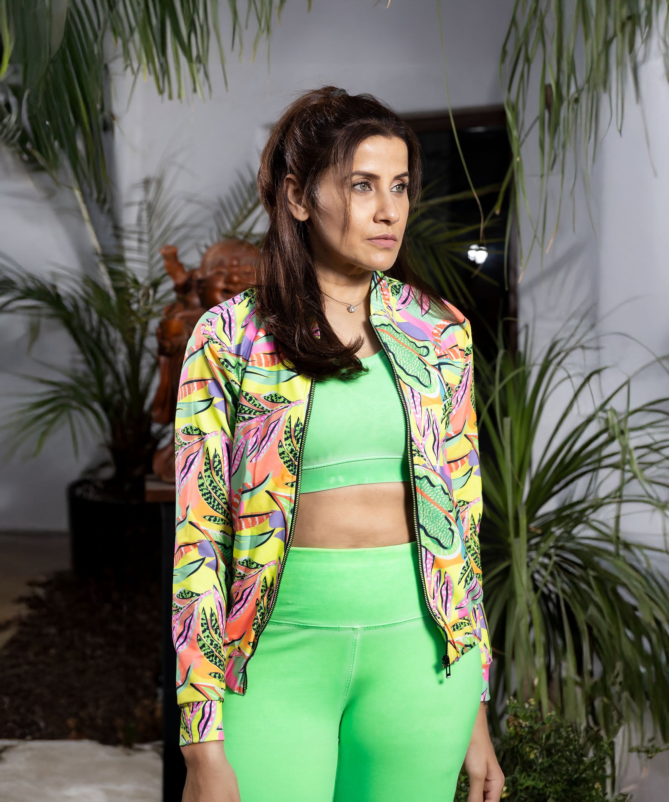 A woman in a bright green outfit and Neon colour Printed Jacket stands among lush plants, gazing to the side.