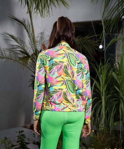 Person with long brown hair, wearing the Neon colour Printed Jacket made from polyblended fabric and bright green pants, stands among plants.