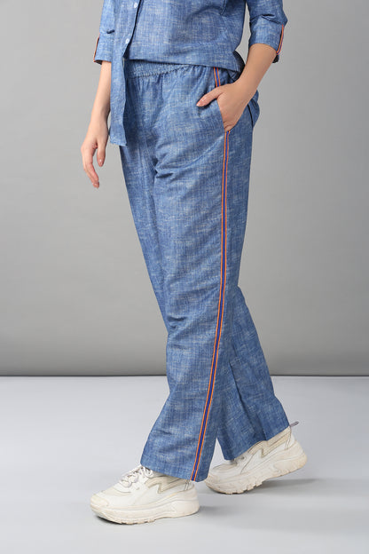 Model wears the Denim Linen Co-Ord Set—blue pants with side stripes and a matching relaxed fit shirt—paired with white sneakers, standing against a gray background.