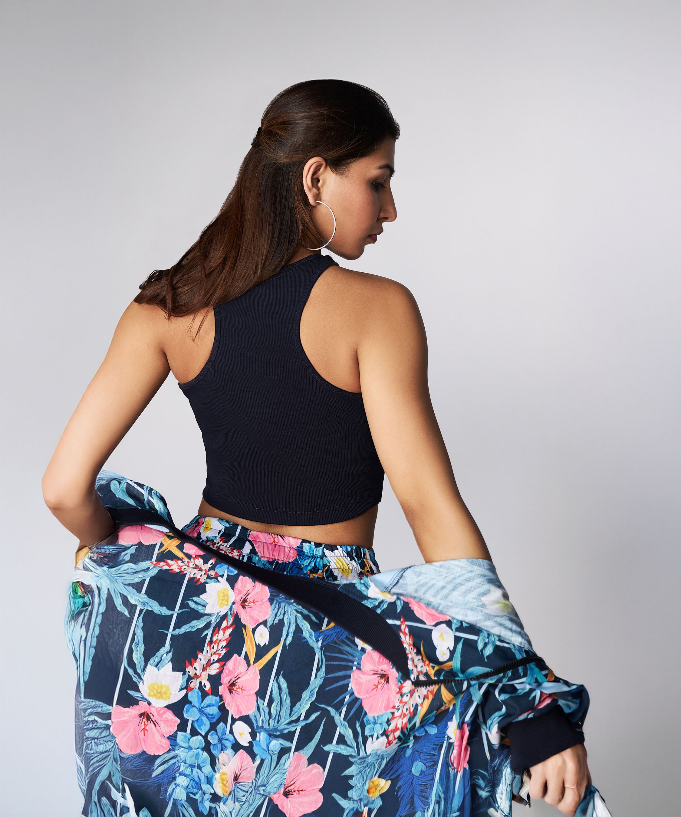 A woman with long brown hair, wearing the Tuna Active Floral Co-ord Set—a black sleeveless top and floral skirt—faces away while holding a matching floral bomber jacket.
