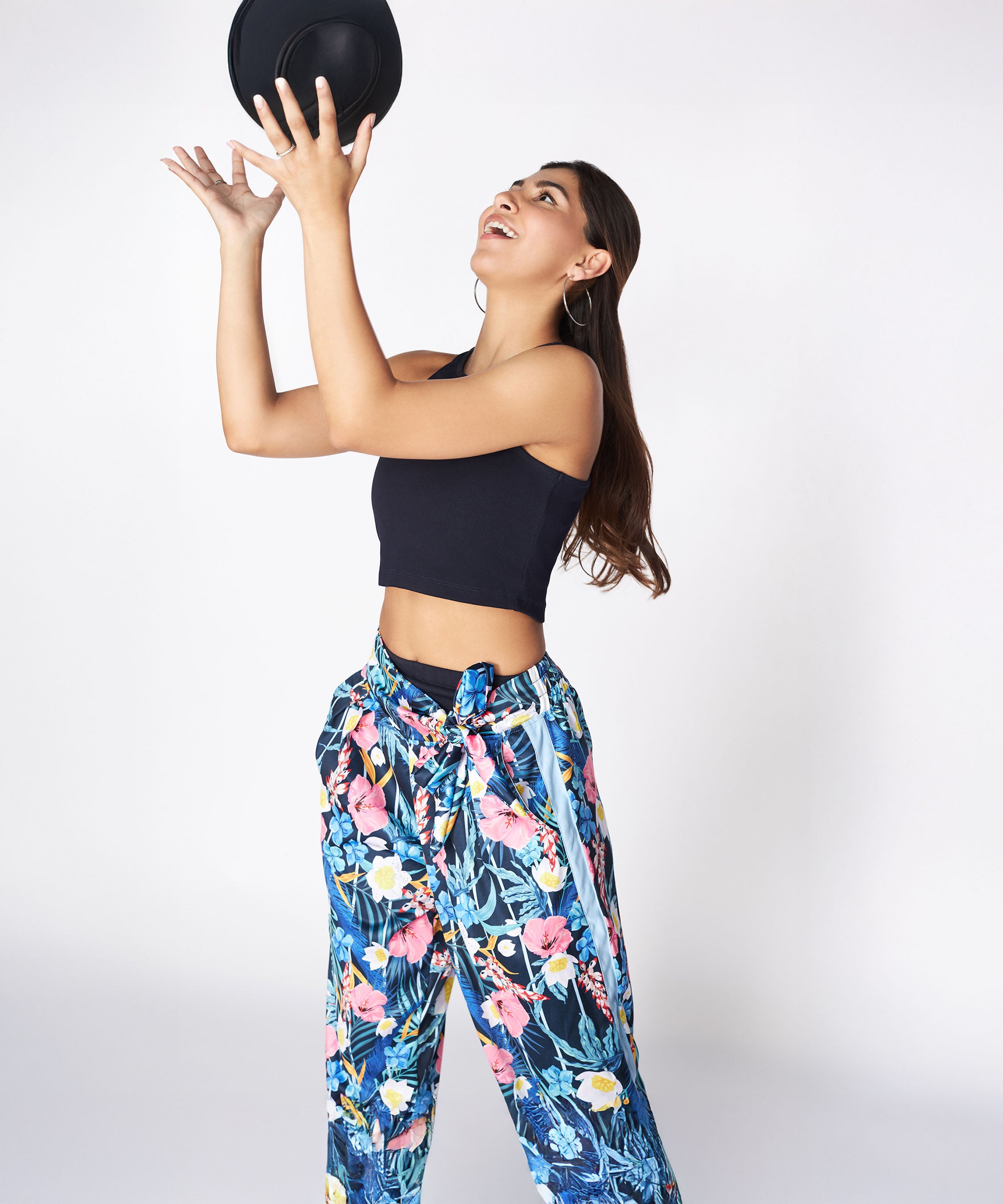 Woman in the Tuna Active Floral Co-ord Set and a black crop top, tossing a black hat in the air while smiling, styled with a floral bomber jacket.