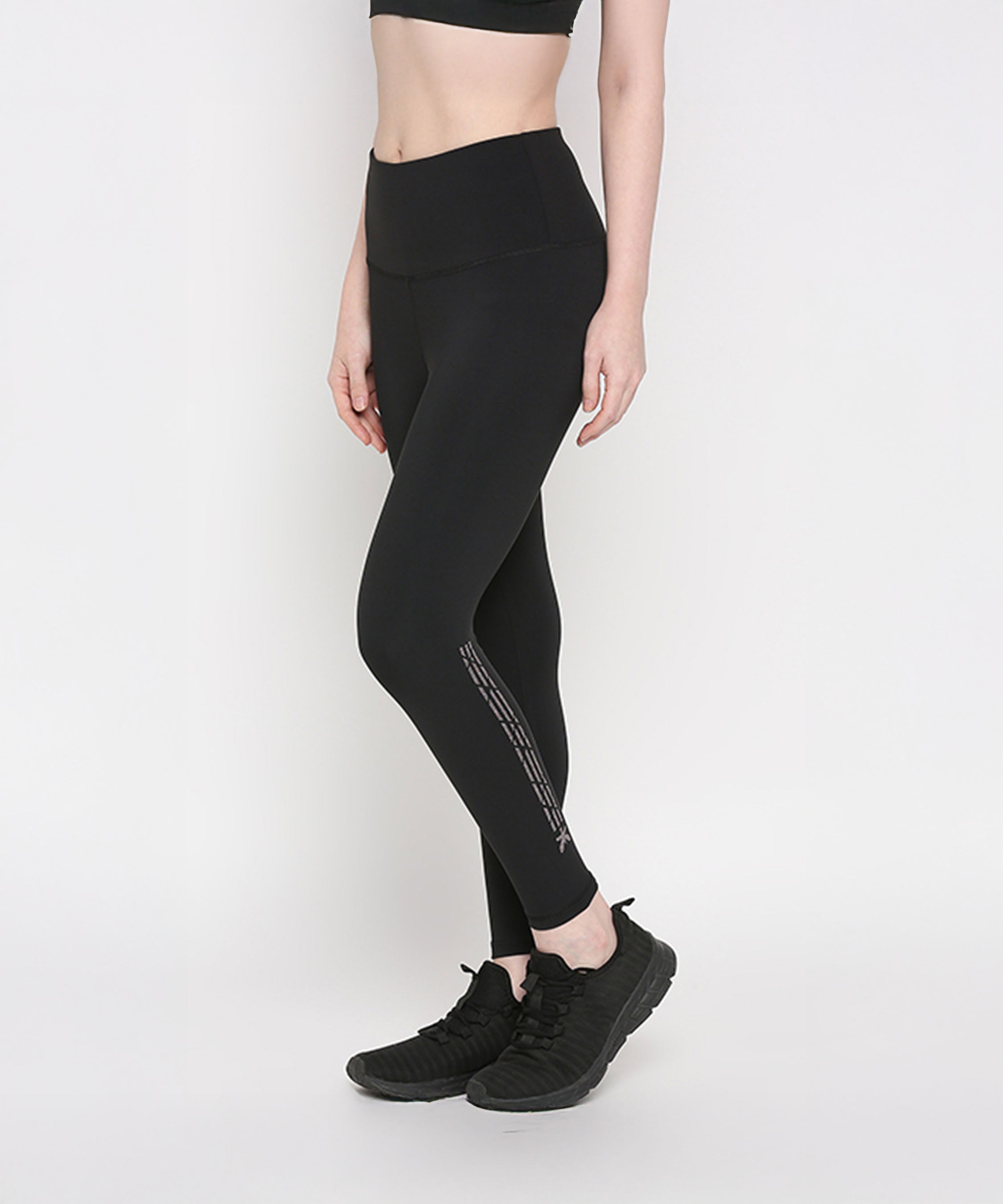 A woman wearing Tuna Active Black Leggings With Print and black sneakers stands against a plain white background, showcasing stylish women’s gym wear.
