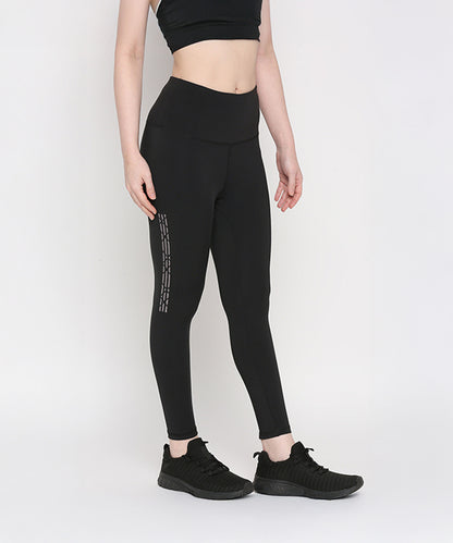 Woman wearing Tuna Active Black Leggings With Print, paired with a black crop top and sneakers, stands against a white background, highlighting stylish women's gym wear.