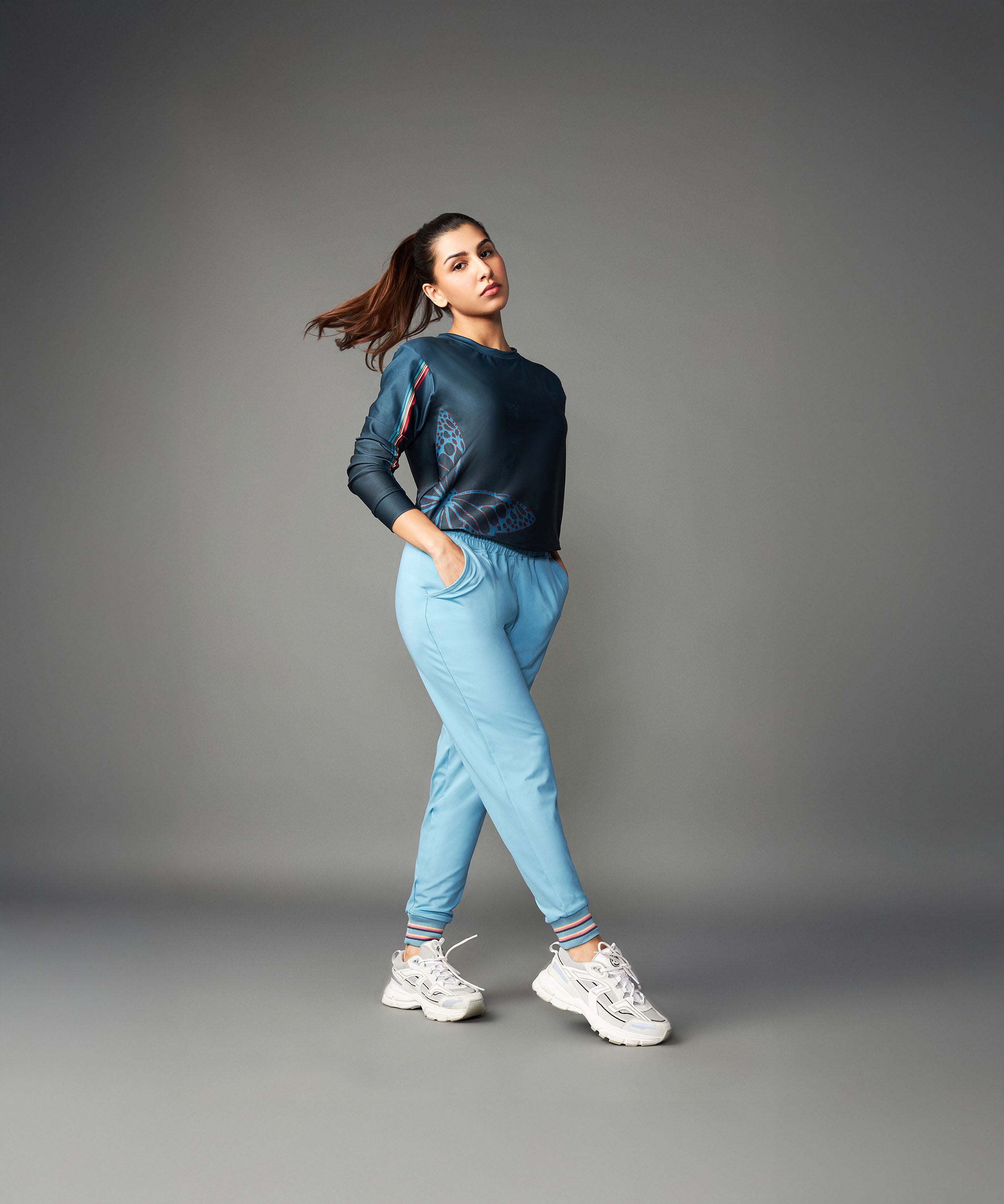 A woman stands confidently against a plain gray background in white sneakers and Tuna Active Blue Joggers, showcasing their comfort.