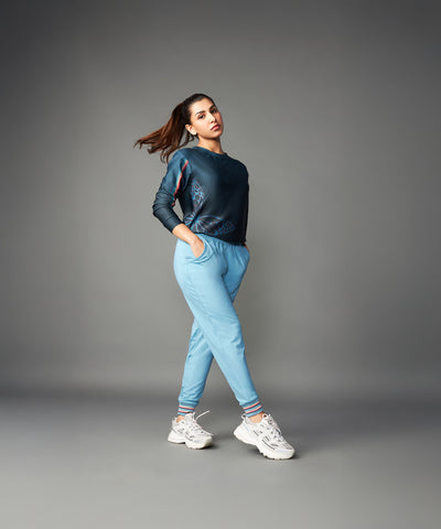 A woman stands confidently against a plain gray background in white sneakers and Tuna Active Blue Joggers, showcasing their comfort.