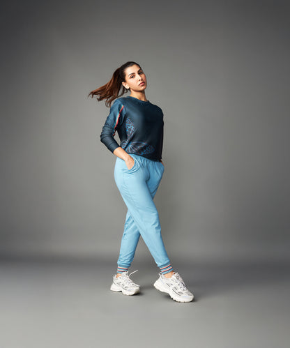 A woman stands confidently against a plain gray background in white sneakers and Tuna Active Blue Joggers, showcasing their comfort.