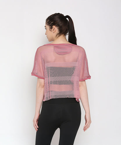 A woman with her hair in a ponytail faces away, wearing the Pink Netted Top in sheer pink over a black sports bra and paired with stylish leggings for a sporty look.