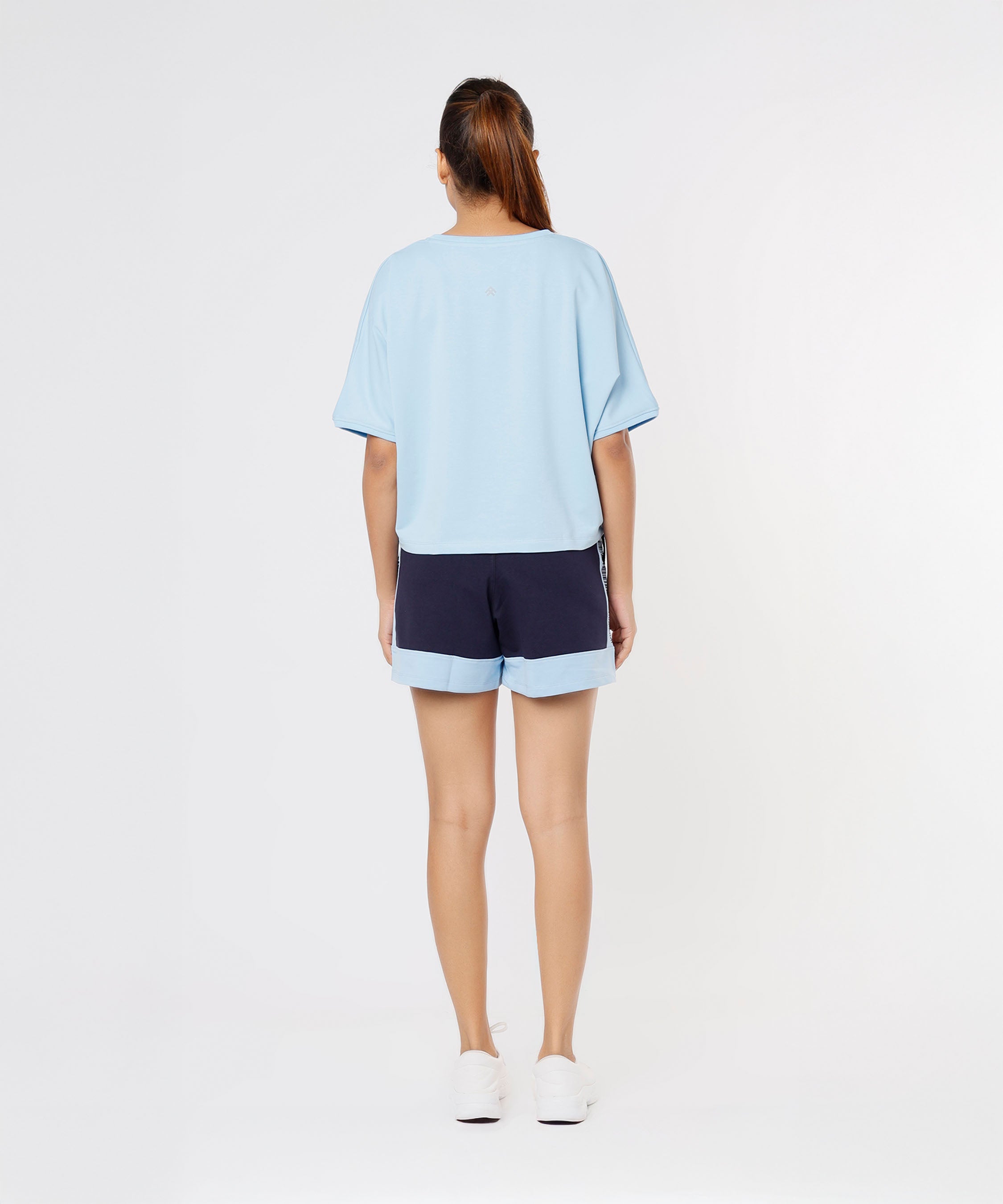 A woman stands facing away, wearing Tuna Active Barcode Printed Blue Shorts as part of a casual co-ord set with a light blue top and white shoes, set against a white background.