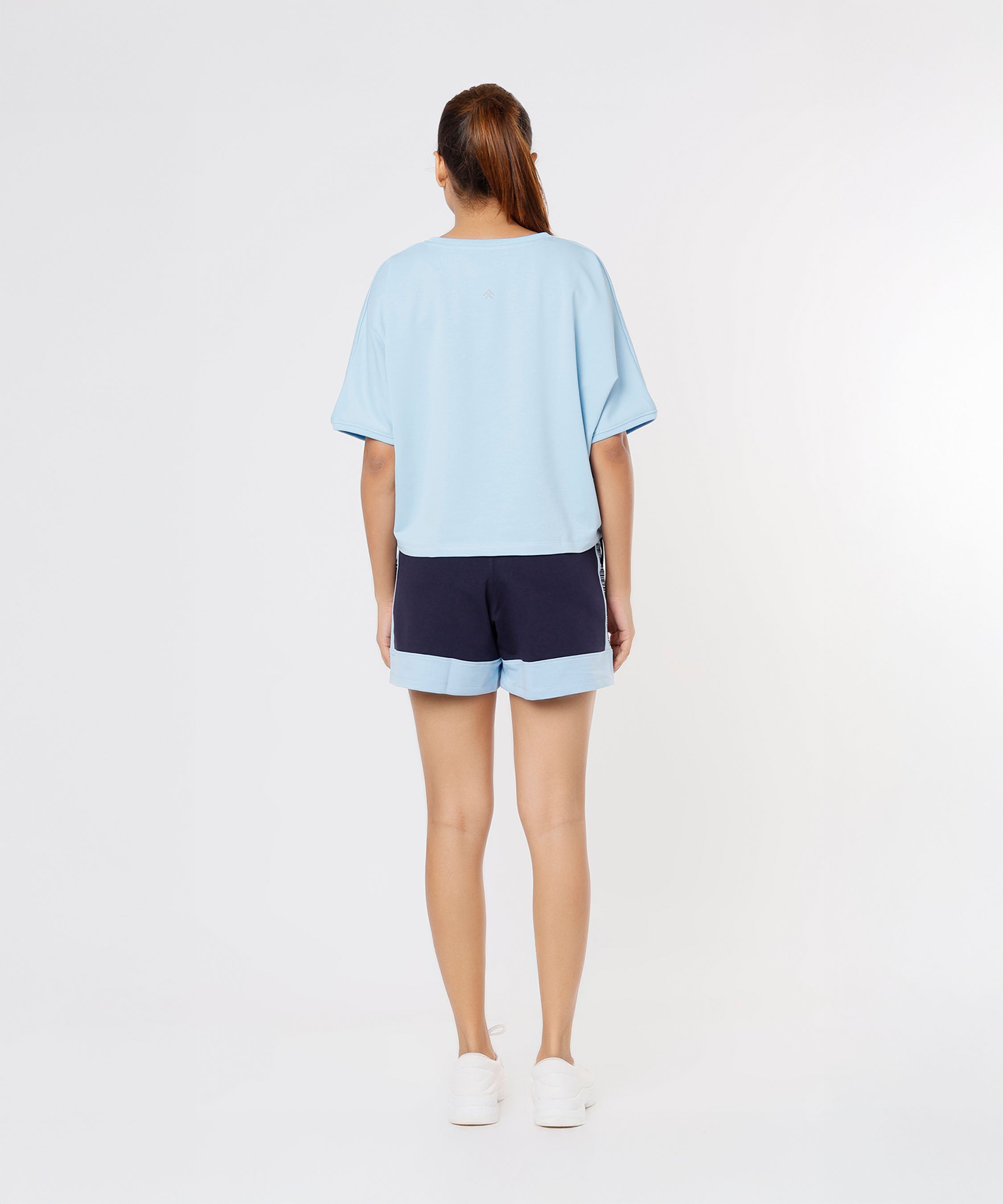 Woman facing away, wearing a light blue cotton-lycra Tuna Active Blue Tee, navy blue shorts, and white shoes against a plain background.