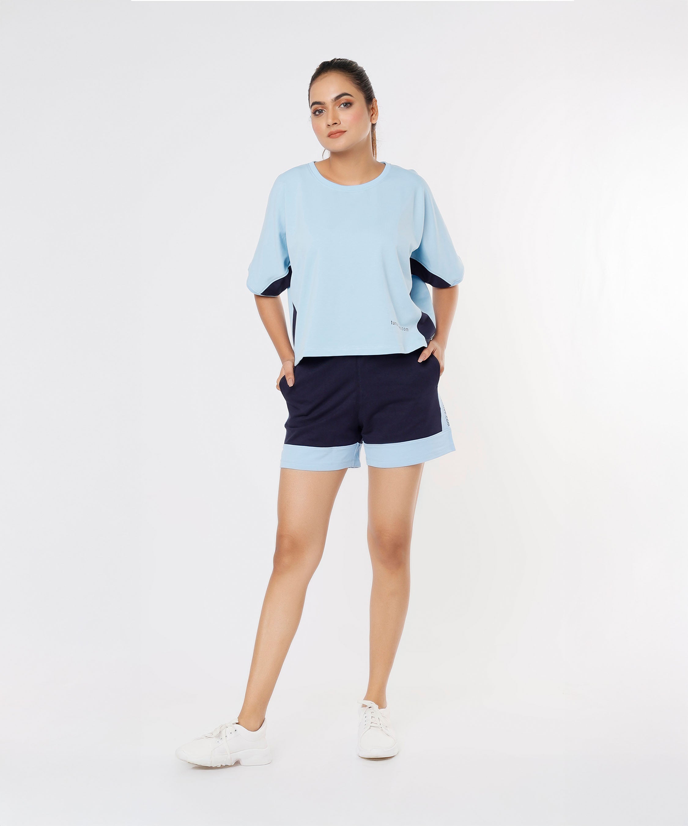 Woman wears the Tuna Active Blue Tee and navy joggers, standing with hands in pockets against a white background.