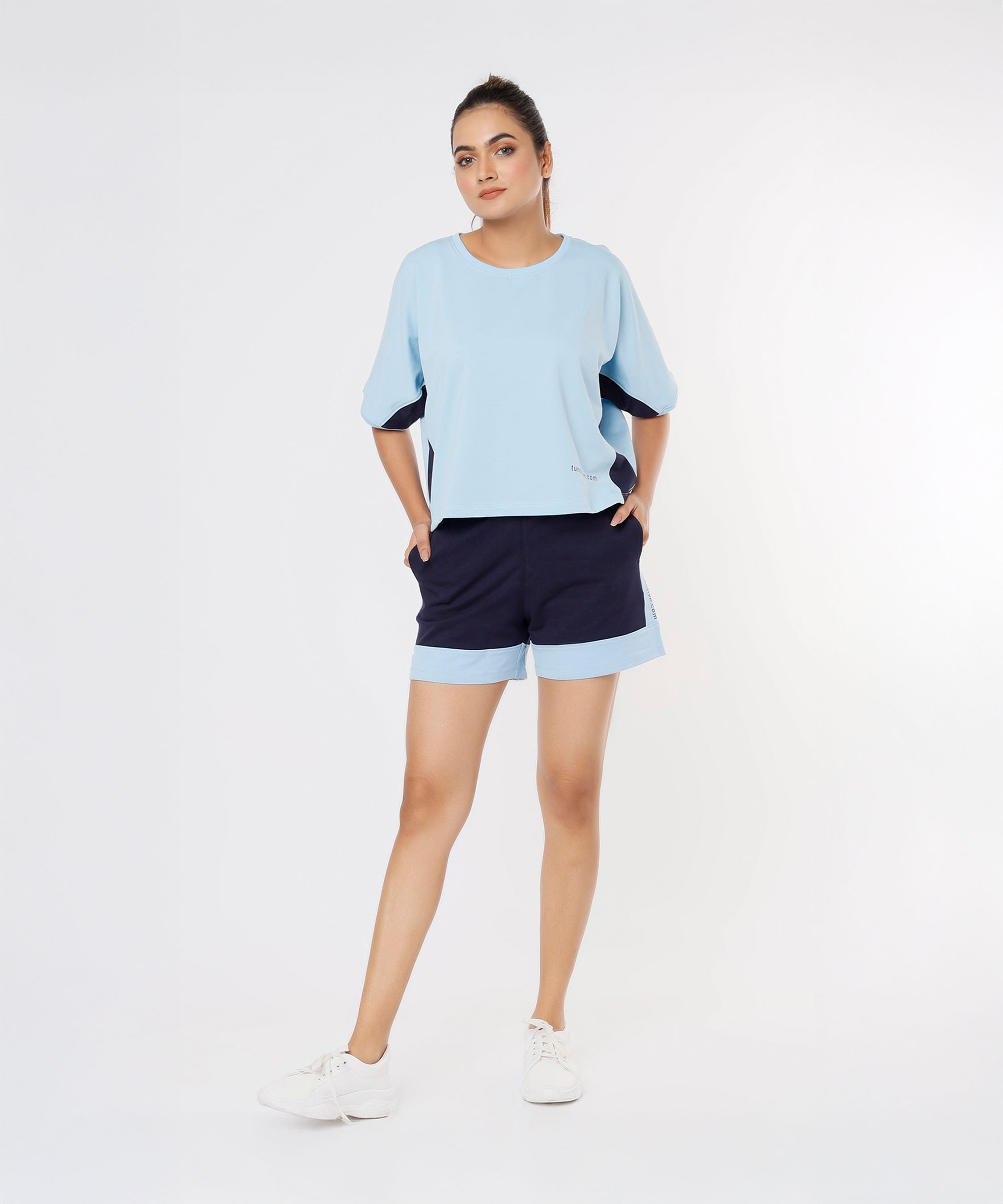 Woman wears Tuna Active Barcode Printed Blue Shorts and a light blue top, standing with hands in pockets against a plain white background.