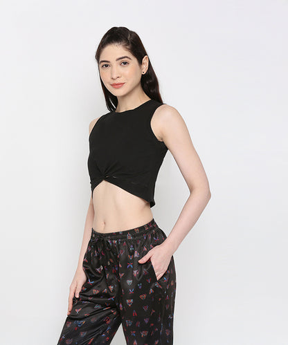 Woman wearing the Black Twisted Crop Top—a black sleeveless, round neck crop top with a twist pattern—paired with printed black pants, stands stylishly with one hand in her pocket against a white background.