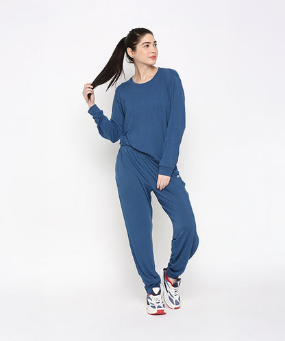 A woman in Atlantic Blue Active Duo loungewear—featuring soft Cotton Modal Joggers and a Blue Racer Back Top—stands against a white background, holding her ponytail and wearing sneakers for ultimate comfort.