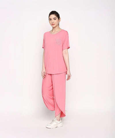 A woman wears the Coral Blissful Tulip Set—coral pink top and matching tulip pants in soft cotton modal—paired with white sneakers, standing against a white background.