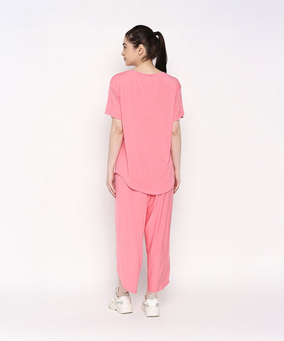 A woman stands facing away against a plain white background, wearing the Coral Blissful Tulip Set— a cotton modal coral pink top and tulip pant loungewear ensemble—paired with white sneakers.