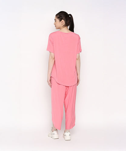 A woman stands facing away against a plain white background, wearing the Coral Blissful Tulip Set— a cotton modal coral pink top and tulip pant loungewear ensemble—paired with white sneakers.