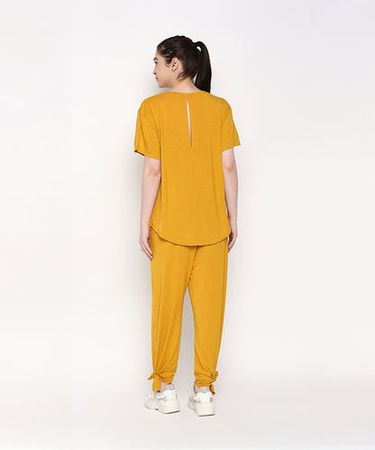 A woman models the Mustard Chic Set—featuring a mustard yellow top and cotton modal trackpant with side knots and a soft elasticated waistband—posed against a white background, facing away.