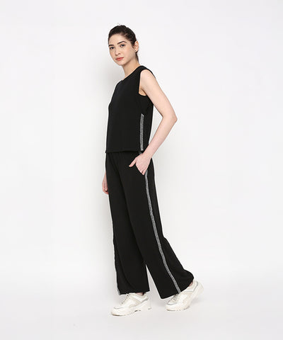 Woman wearing the Comfy Black Set with Side Sparkle, a black sleeveless top and loose fit track pant featuring side stripes, crafted from comfortable cotton lycra, standing against a white background.