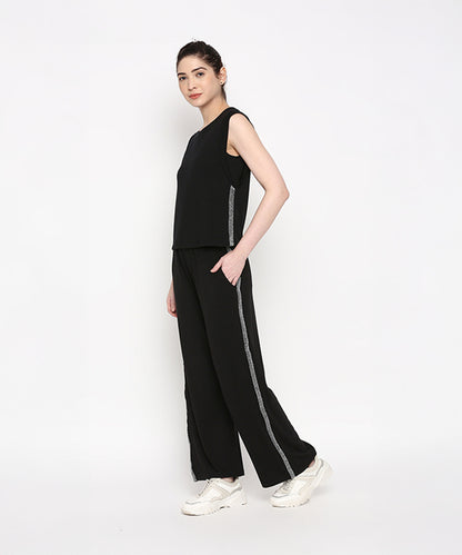 Woman wearing the Comfy Black Set with Side Sparkle, a black sleeveless top and loose fit track pant featuring side stripes, crafted from comfortable cotton lycra, standing against a white background.