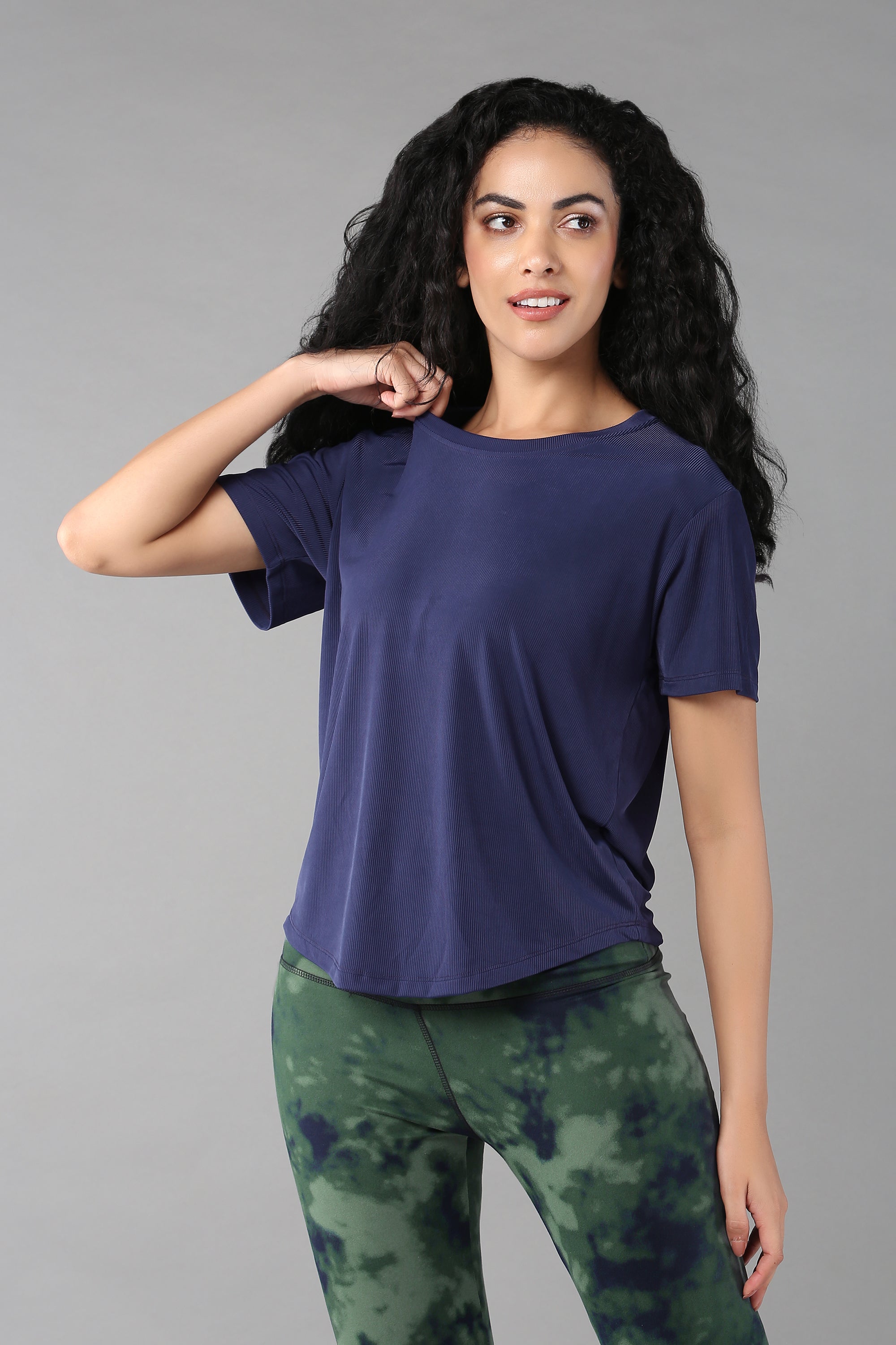 Woman with long curly hair wears the Functional - Blue Soft T Shirt and green tie-dye leggings, standing against a gray background—an effortlessly stylish and versatile look for any adventure.