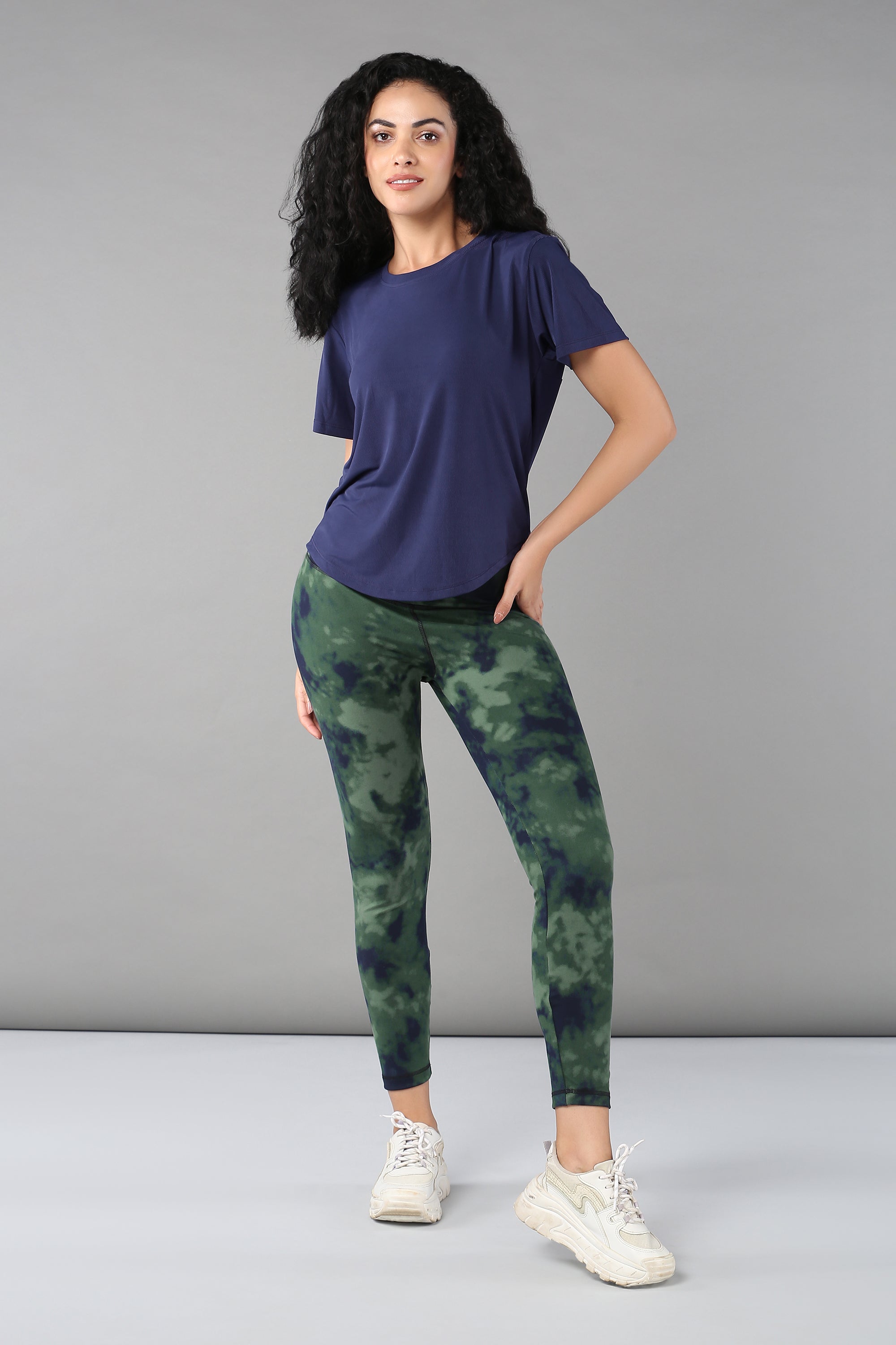 Woman in a Functional - Blue Soft T Shirt, green tie-dye leggings, and white sneakers poses against a gray background—an ideal versatile look for stylish adventures.