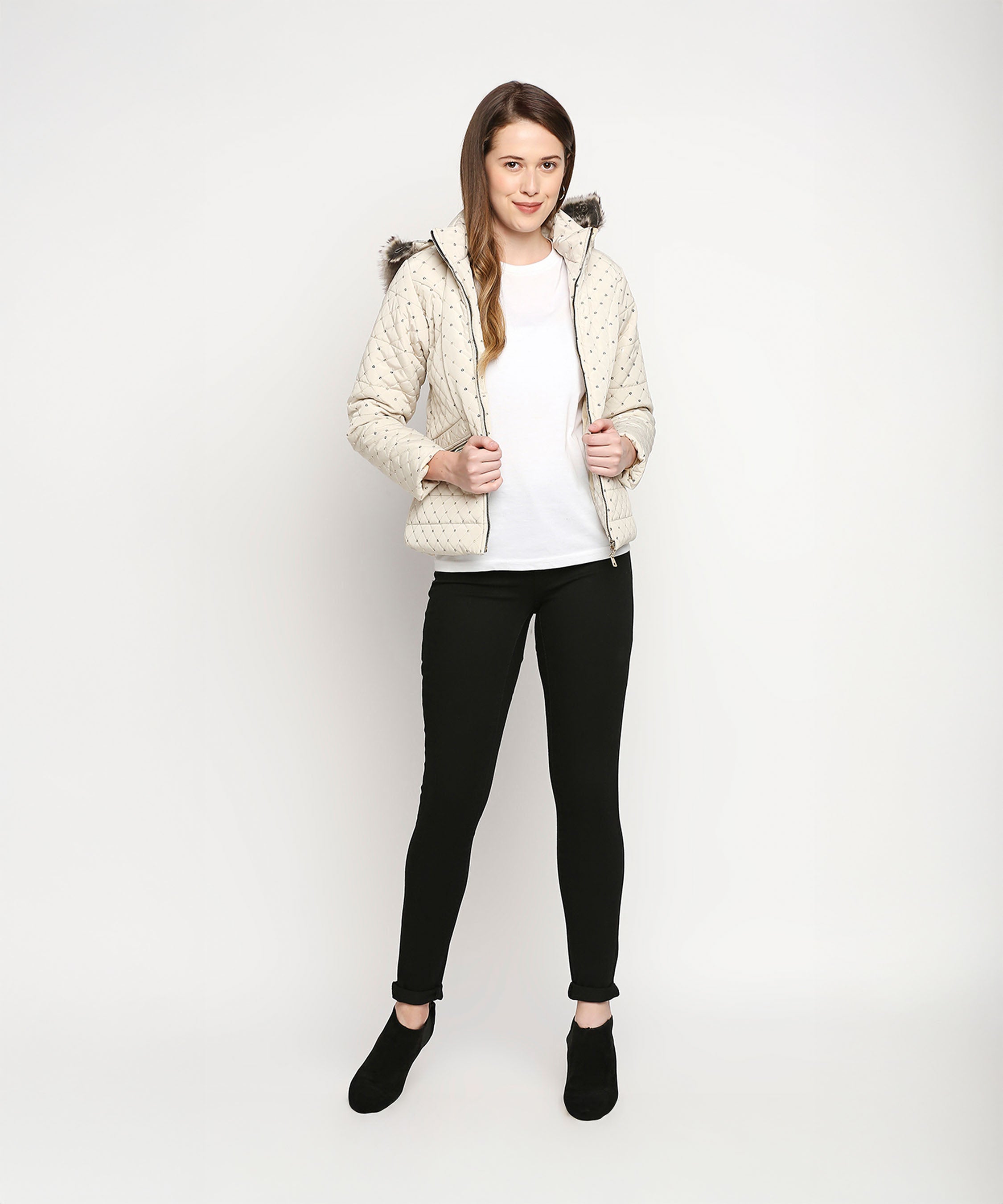A woman wearing an Off White Mid Length High Collar Puffer Jacket With Detachable Fur Hoodie, a white shirt, black pants, and black shoes stands against a white background.