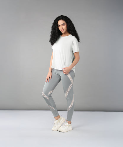 Woman with long curly hair wears the Grey Top with Patched Legging, made from super soft, moisture-wicking fabric, paired with sneakers and poses against a plain gray background.