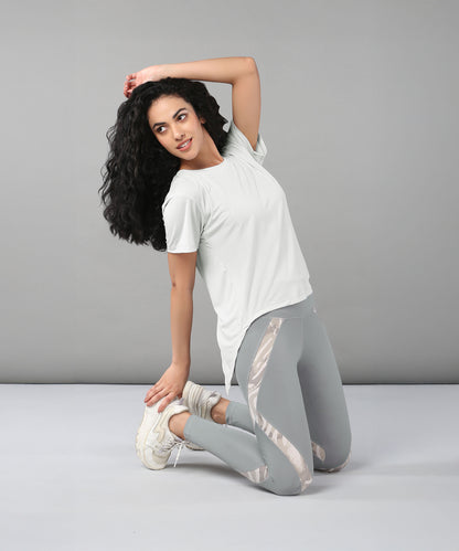 A woman in a Grey Top with Patched Legging kneels and stretches on a plain gray background. The super soft, moisture-wicking fabric of her activewear delivers comfort and keeps her cool throughout her workout.