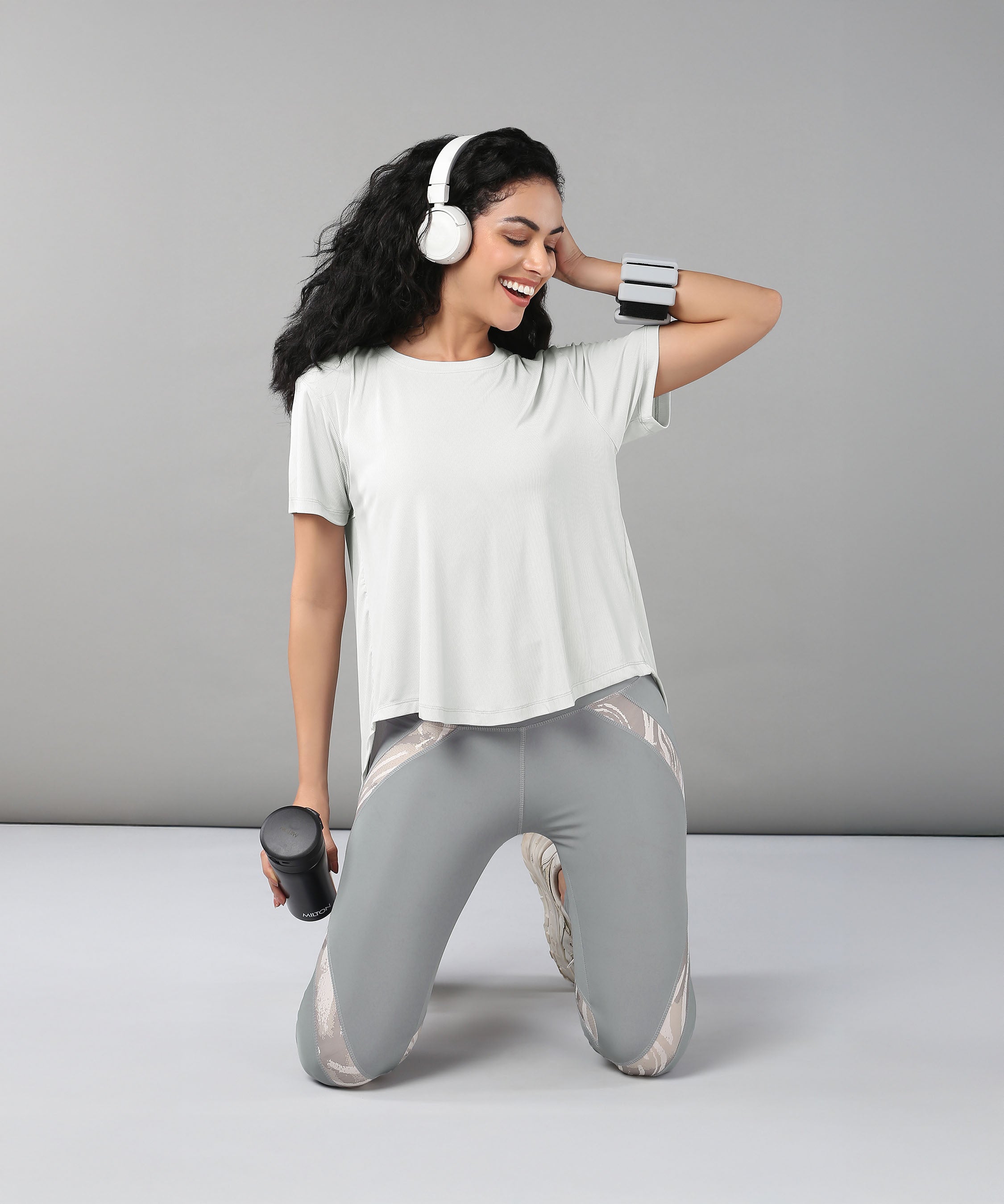 Smiling woman kneeling with headphones and holding a water bottle, dressed in the Grey Top with Patched Legging—workout clothes made from super soft fabric for all-day comfort.